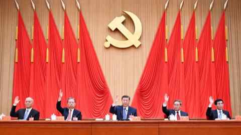 Five Asian men sit behind the stage and raise their hands, while they are covered by ten red flags and a golden hammer and sickle.